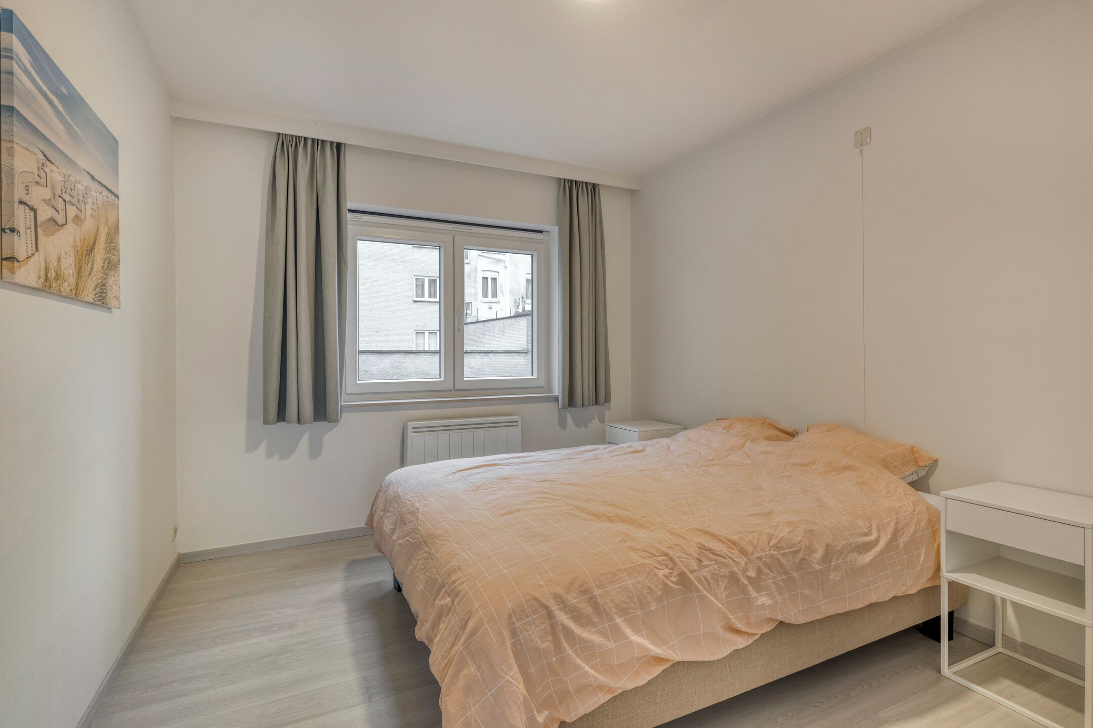 A bedroom with a bed, a window with curtains, a radiator, and a wall-mounted shelf.