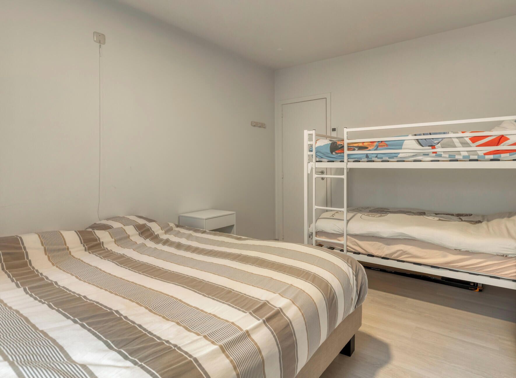A single bed with a striped bedspread, a white nightstand, and a bunk bed with a colorful mattress.