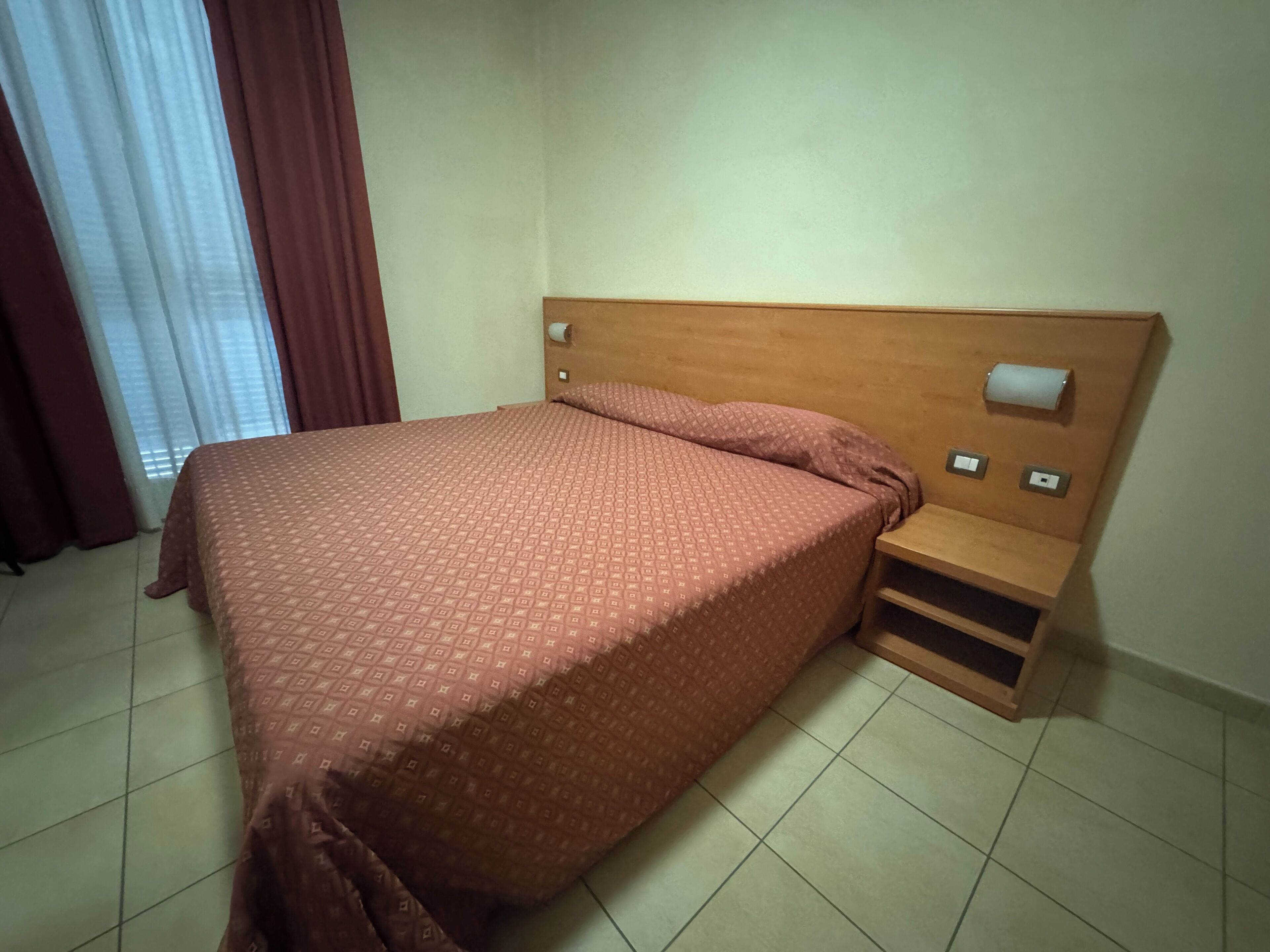 Una cama individual con una colcha estampada, un cabecero de madera, una mesita de noche con una lámpara y una ventana con cortinas.