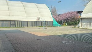 Ein gepflasterter Bereich mit einem Basketballkorb, einer grünen Spielanlage und einem überdachten Gebäude.