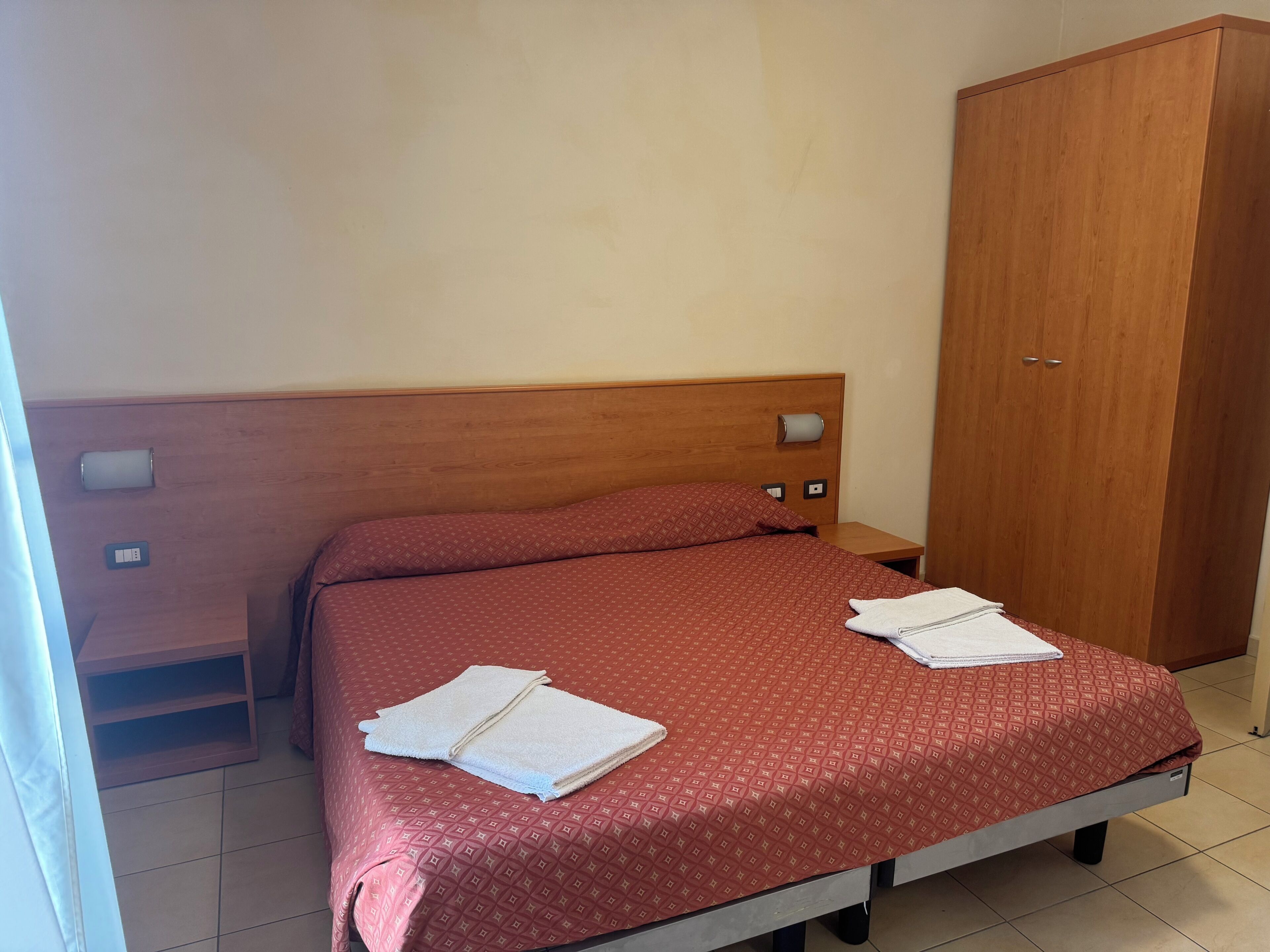 Una cama individual con colcha estampada roja, cabecera de madera, mesita de noche, armario de madera y un enchufe en la pared.