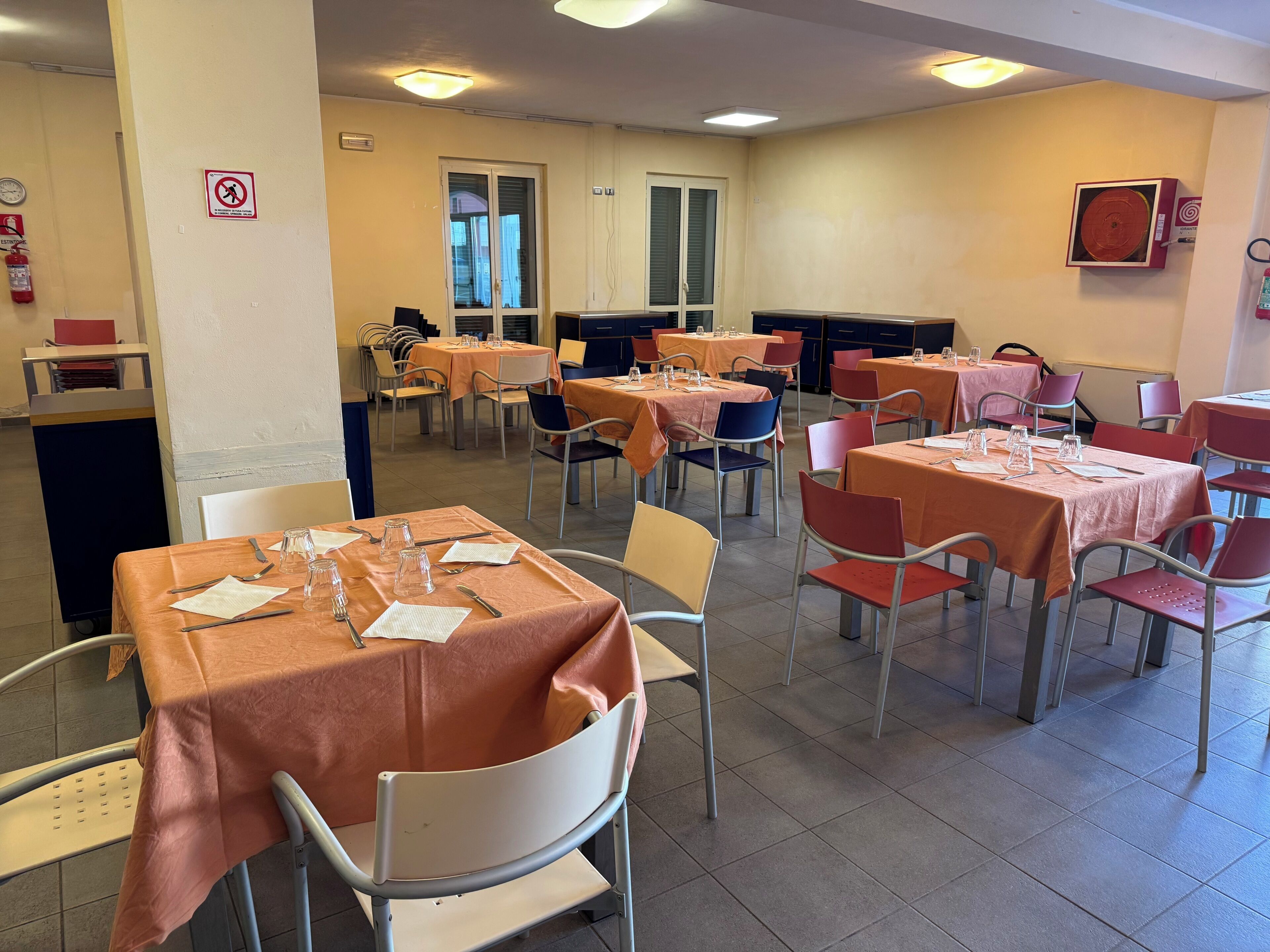Un comedor con mesas dispuestas para el servicio, sillas y un extintor en la pared.