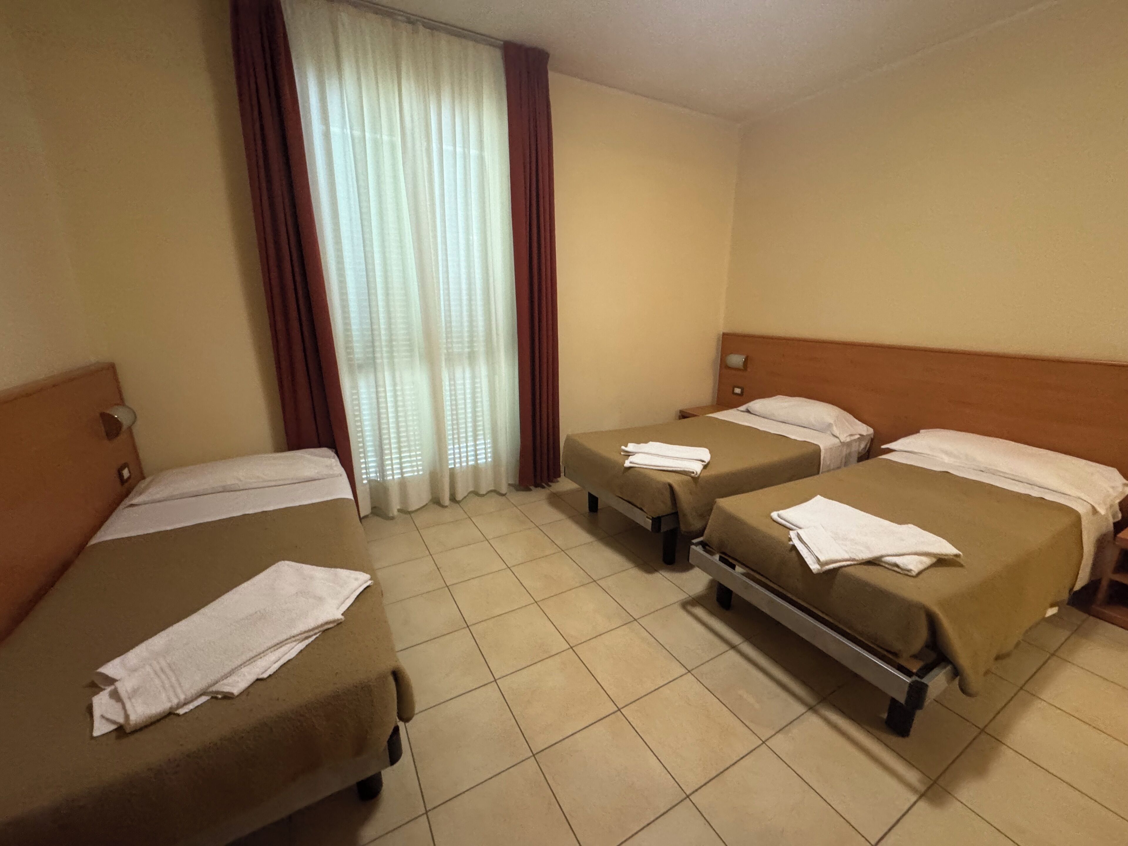 A hotel room with three single beds, a window with curtains, and a tiled floor.