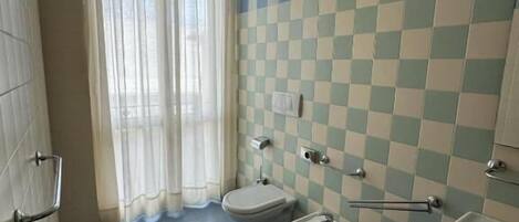 Un baño con pared de baldosas a cuadros, un inodoro, bidés, un lavabo y un cubo de basura.