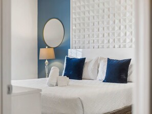 Una cama bien tendida con almohadas blancas y azules, un espejo redondo en la pared y un cabecero estampado.