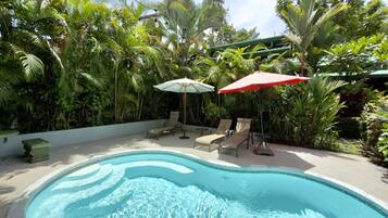 En swimmingpool med liggestole og parasoller, omgivet af frodig, tropisk vegetation.
