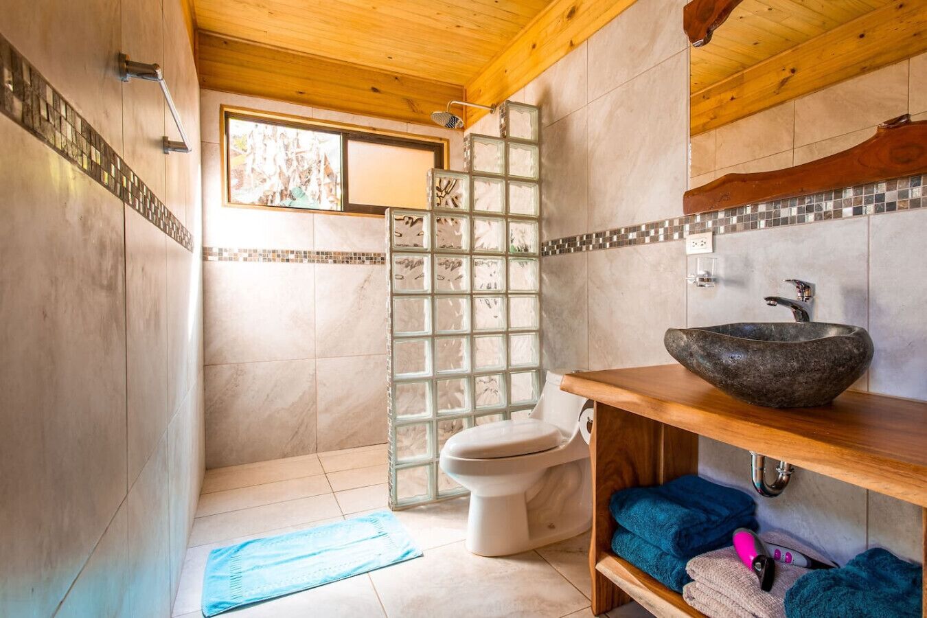Un baño con techo de madera, cabina de ducha de vidrio, inodoro y lavamanos con cuenca de piedra.