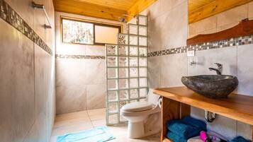 Un baño con techo de madera, cabina de ducha de vidrio, inodoro y lavamanos con cuenca de piedra.