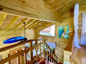 Interior de una cabaña de madera con una escalera de madera, un objeto azul en el techo, un cuadro de un loro y una ventana con vistas a un edificio rojo.