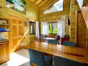 Interior de una cabaña de madera con una mesa de comedor, sillas, una zona de cocina y una ventana con cortinas.