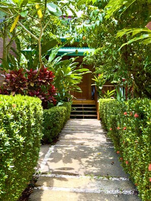 A pathway leading to a wooden structure with a green roof, surrounded by well-maintained hedges and various plants. - Tropical Eco Cabin near Beach & Park (Manuel Antonio)