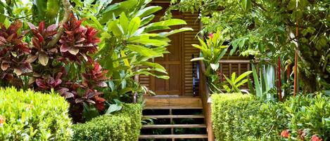A wooden staircase leading to a building with a green roof, surrounded by lush greenery and various plants.