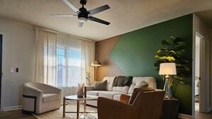 Ett modernt vardagsrum med en soffa, en fåtölj och ett soffbord. Det finns en takfläkt, en lampa och en växt.