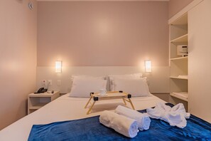 Een hotelkamer met een net opgemaakt bed, een nachtkastje met een telefoon en afstandsbediening, een bijzettafeltje met een kopje en afstandsbediening, en een garderobe met opgevouwen kleding.