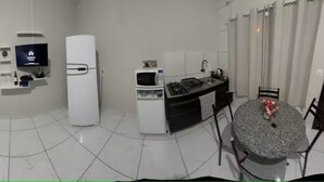 Un soggiorno moderno con un divano grigio, una TV a schermo piatto montata a parete, un frigorifero bianco, un angolo cottura con forno a microonde e fornelli, e un tavolo da pranzo con sedie.