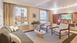 Un salon moderne avec un canapé, des fauteuils, une table basse et un téléviseur fixé à un mur en bois. De grandes fenêtres offrent une vue sur un bâtiment situé à l’extérieur.