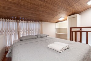 A bedroom with a wooden ceiling, a bed with a grey quilt, two pillows, and a white towel on the bed. There is a window with curtains and a built-in shelf on the wall. - Solar Condominium Agua Pipa by Carpediem (Tibau do Sul)