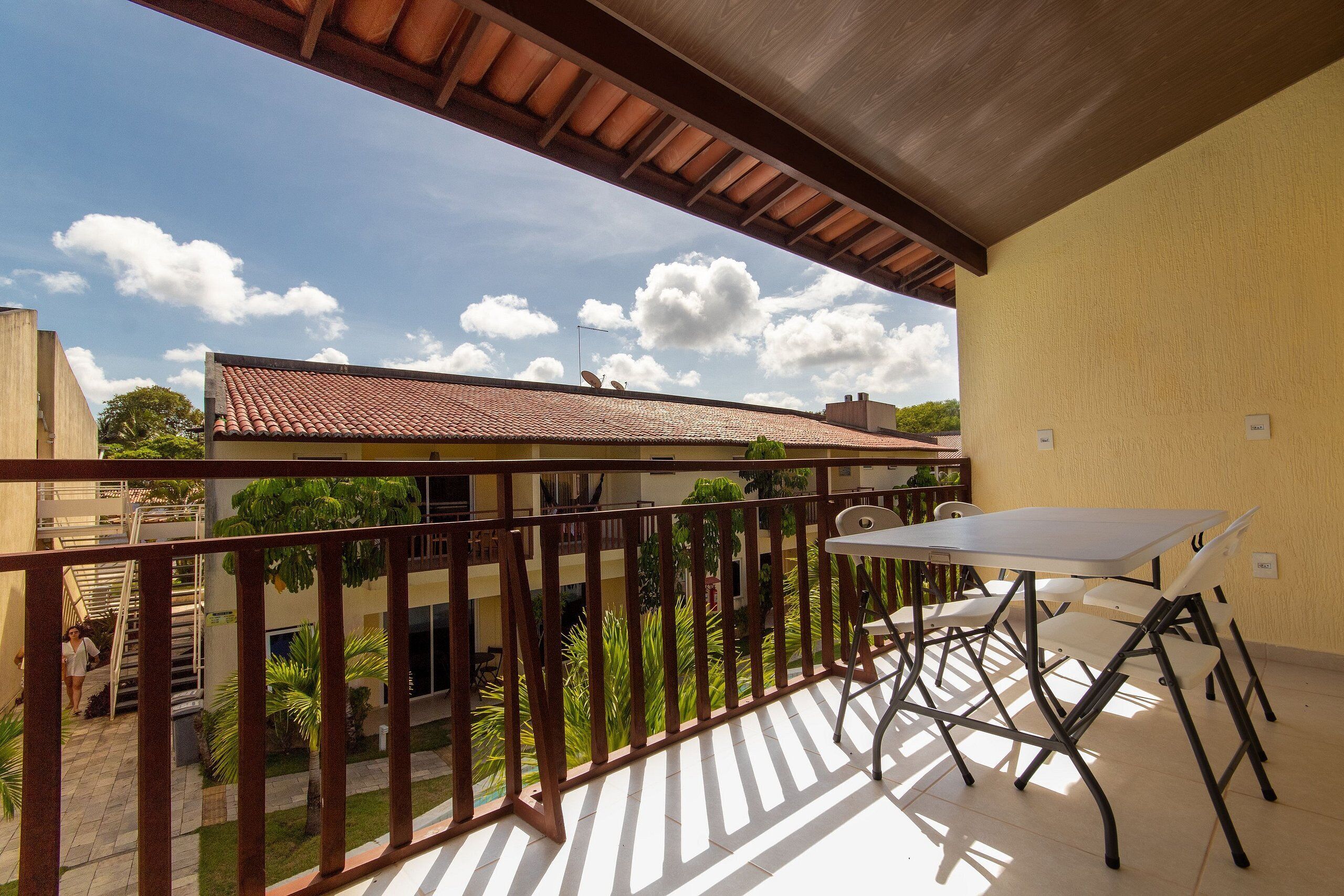 A balcony with a table and chairs, a view of a building with a tiled roof, and a clear sky with clouds.