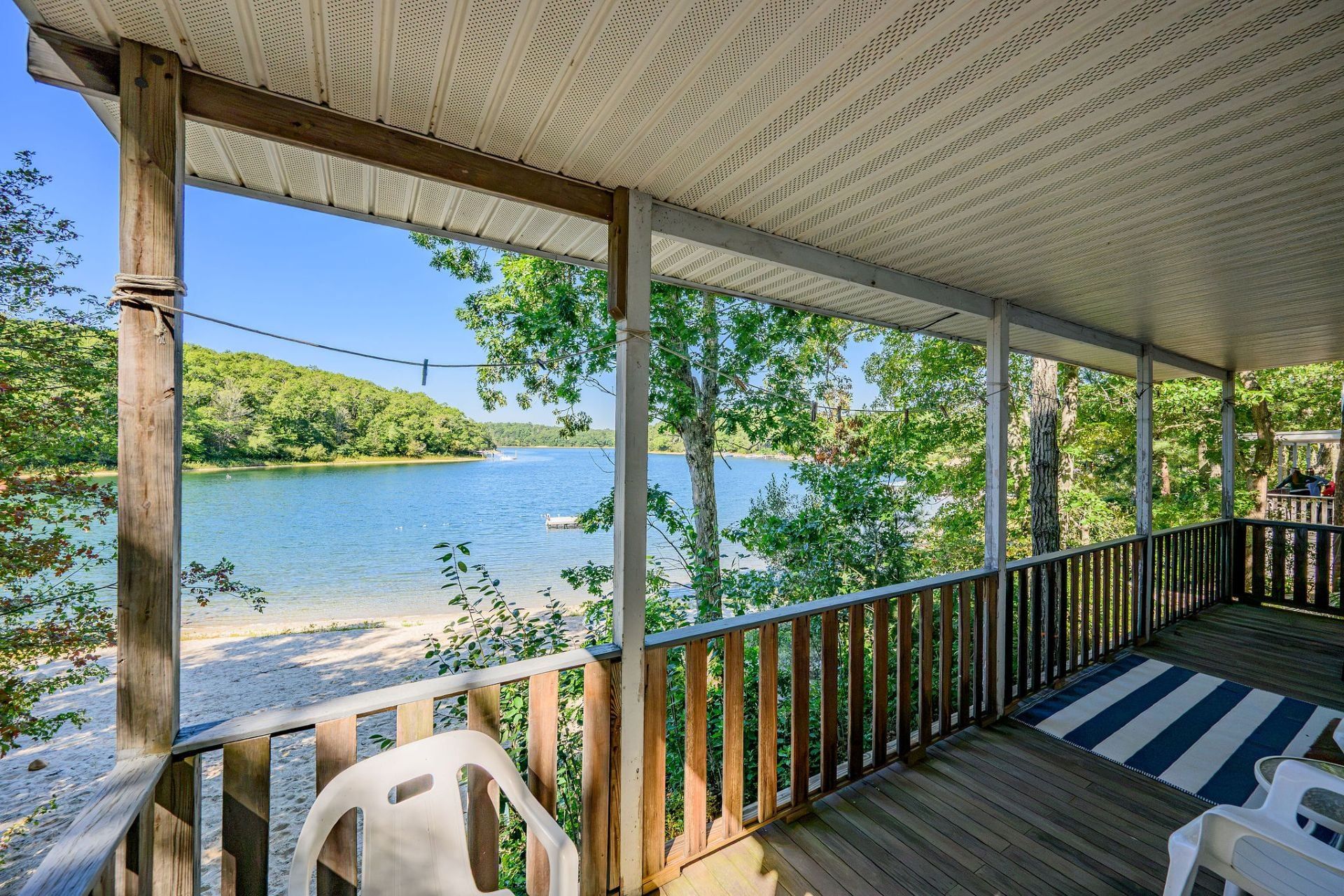 A covered deck with a view of a lake, wooden railings, and a striped rug.