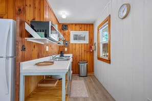 A compact kitchen with wooden paneling, a white countertop, a sink, a stove, and a trash can. - Peaceful Waterfront Cottage with Pond Views (Sandwich)