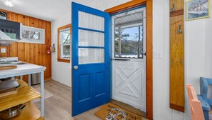 Una habitación con puerta azul, paredes de madera, una zona de cocina con estufa y ventana con vista a árboles.