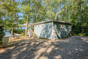 Eine kleine Holzhütte mit Veranda, Picknicktisch und Grill, umgeben von Bäumen und einem Gewässer.