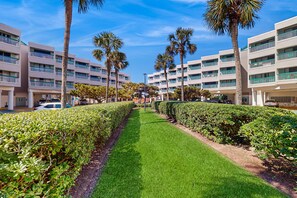 A hotel with a central pathway flanked by neatly trimmed hedges and palm trees, leading to a building with multiple balconies. - Centrally located, cozy condo with shared pool & hot tub - steps to sand (Galveston)