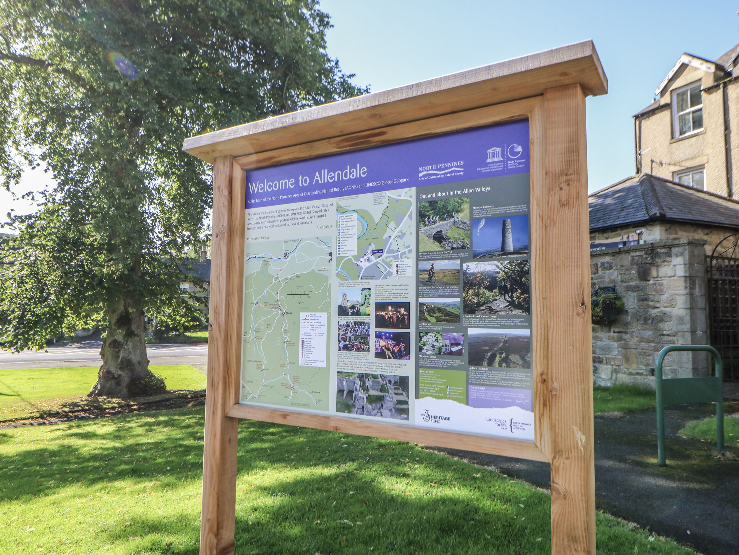 A wooden information board with a map and photos, surrounded by grass and a tree.