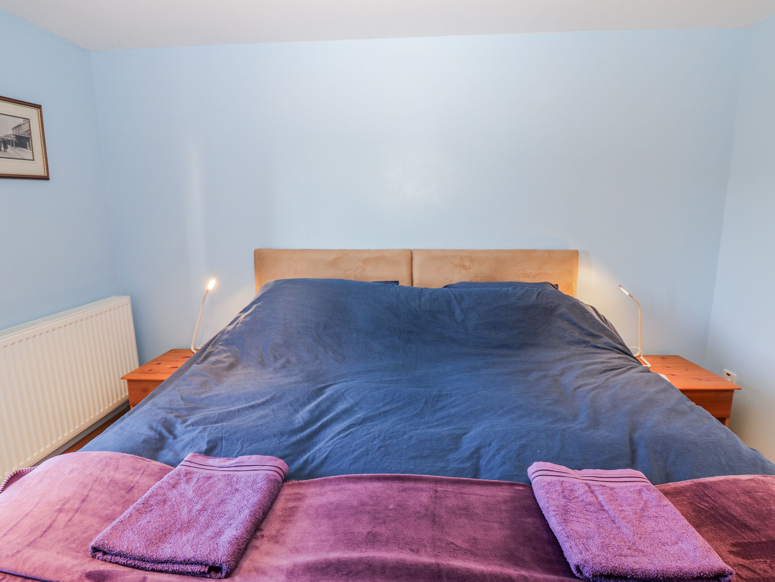 Une chambre à coucher comprenant un lit, deux tables de chevet, une commode, un tableau encadré au mur et un radiateur.