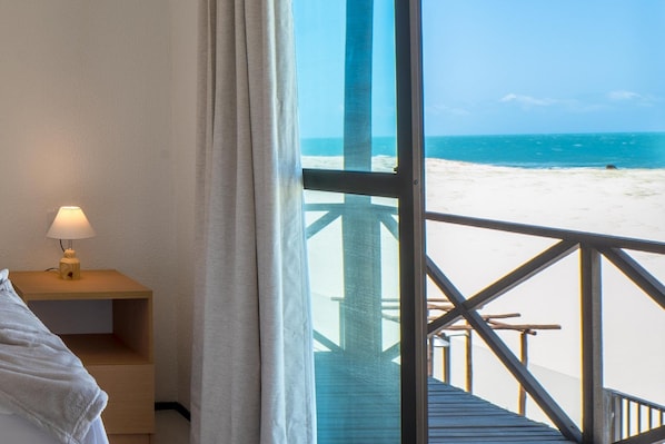 A room with a bed, a nightstand with a lamp, and a large window overlooking a beach.