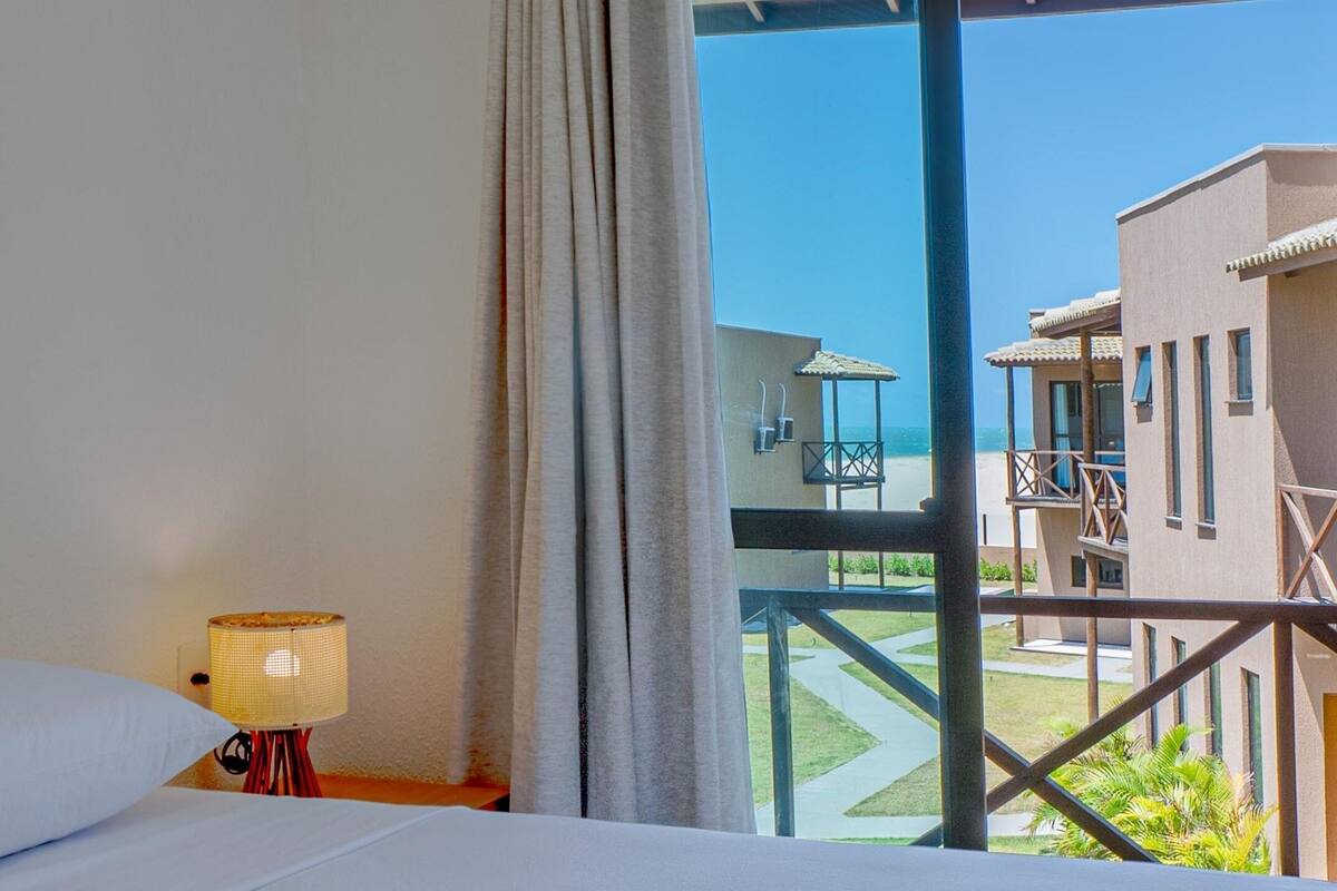 Une chambre d’hôtel avec un lit, une table de chevet avec une lampe, et une vue sur la piscine et les bâtiments environnants.