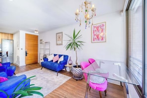 Ein modernes Wohnzimmer mit einem blauen Sofa, einem pinken Sessel und einem Glastisch. Eine Topfpflanze und ein Kronleuchter sind ebenfalls vorhanden.