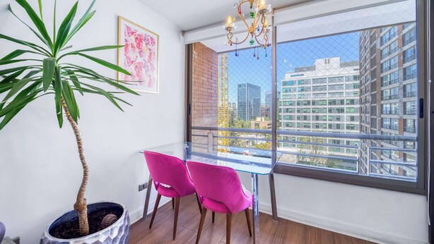 A balcony with a potted plant, two pink chairs, a glass table, and a view of city buildings. - Blue Pink Home - Wifi & Gym near Escuela Militar (Las Condes)