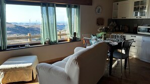 Una habitación con una mesa de comedor, sillas, un sofá, un ventanal con cortinas, vistas al campo y una zona de cocina.