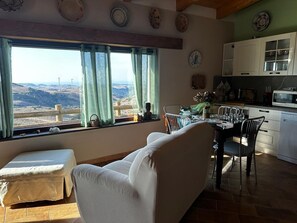 Une pièce comprenant une table à manger, des chaises, un canapé, une fenêtre avec des rideaux, une vue sur la campagne et un coin cuisine.