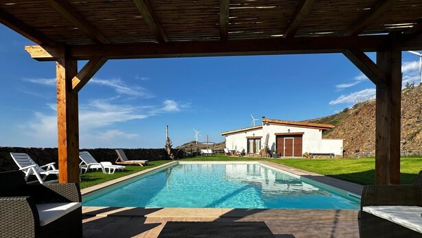 Una piscina con sillones de descanso, una pérgola y un edificio con aerogeneradores al fondo.