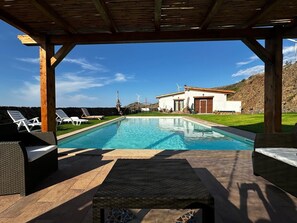 Une piscine avec des chaises longues, une pergola et, au fond, un bâtiment surmonté d’éoliennes.