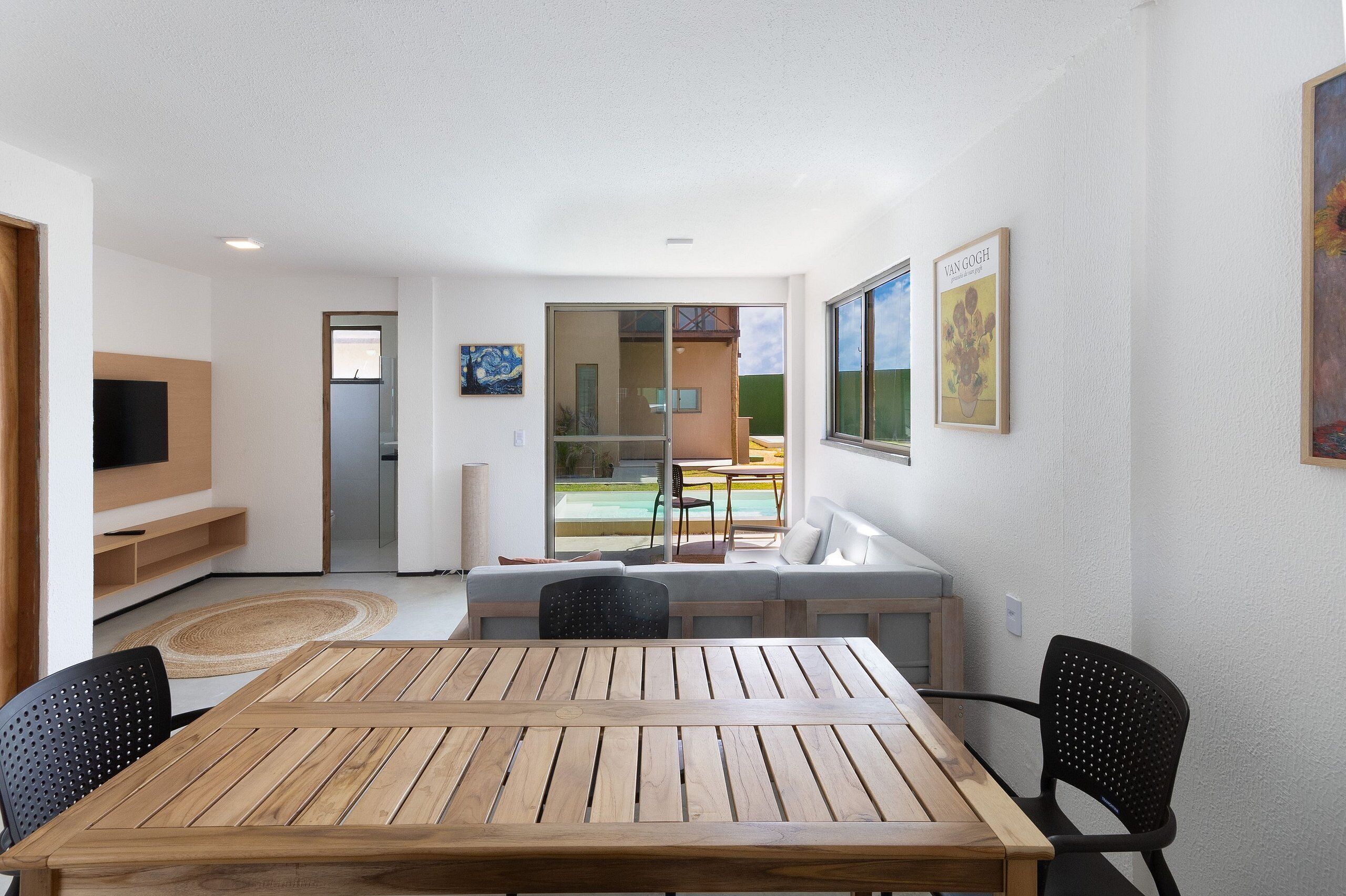 Una sala de estar moderna con una mesa y sillas de madera, un sofá y una televisión. Posee un ventanal corredero que da a un balcón con piscina.