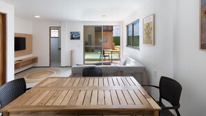 Ein modernes Wohnzimmer mit Holztisch und -stühlen, einer Couch und einem Fernseher. Eine Schiebetür aus Glas führt zu einem Balkon mit einem Pool.