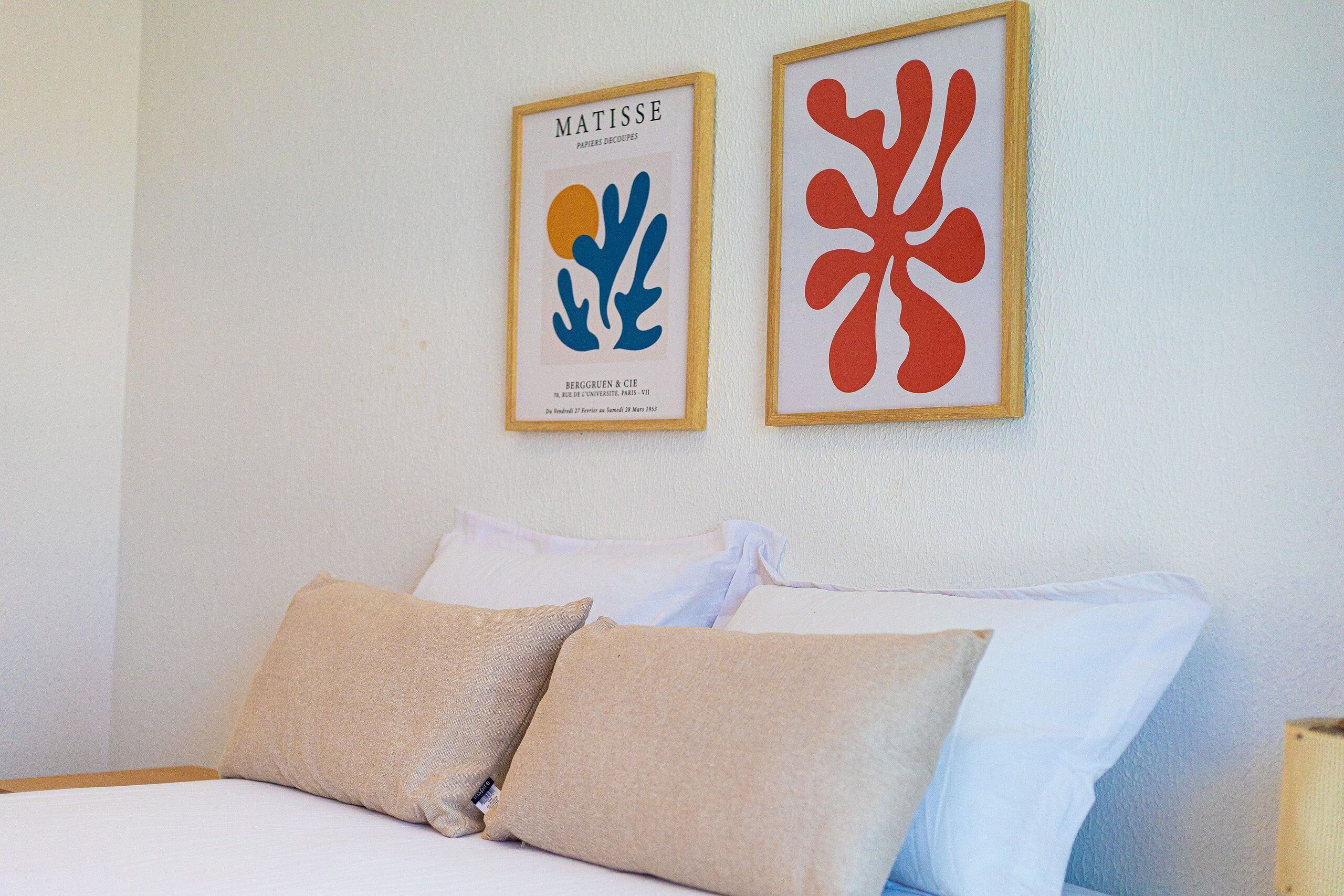 A neatly made bed with beige and white pillows, two framed artworks on the wall above it, and a plain white wall.