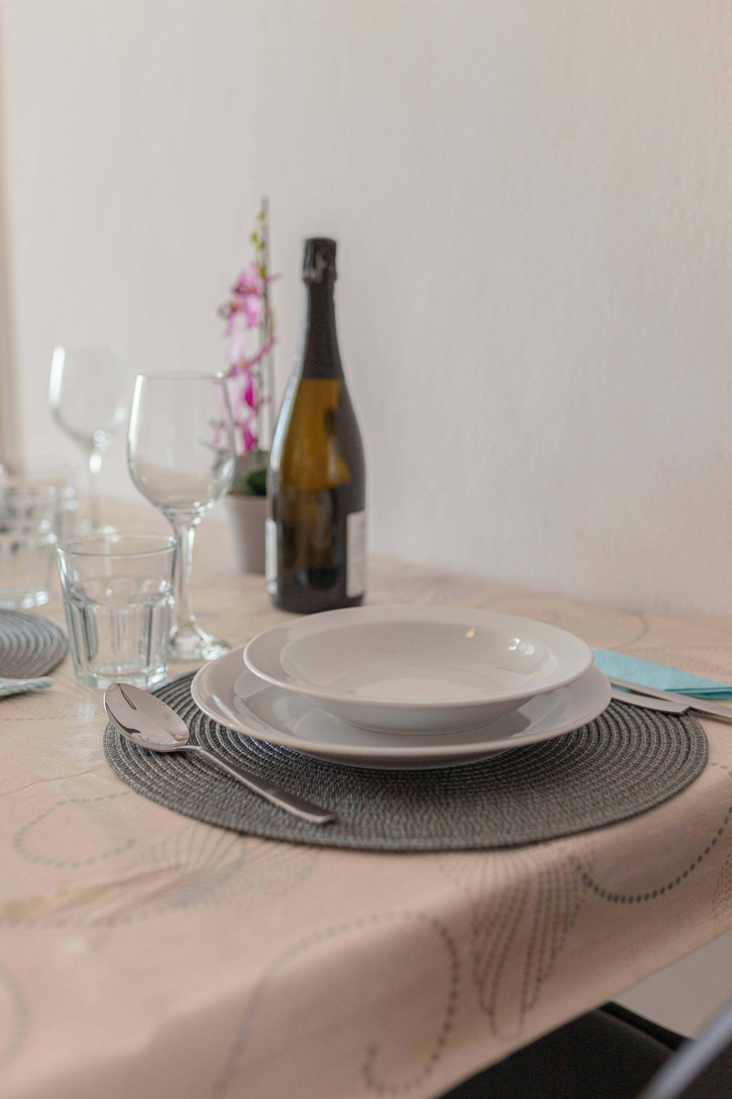 Una mesa de comedor preparada con platos, vasos, cubiertos, una botella de vino y un jarrón con flores.