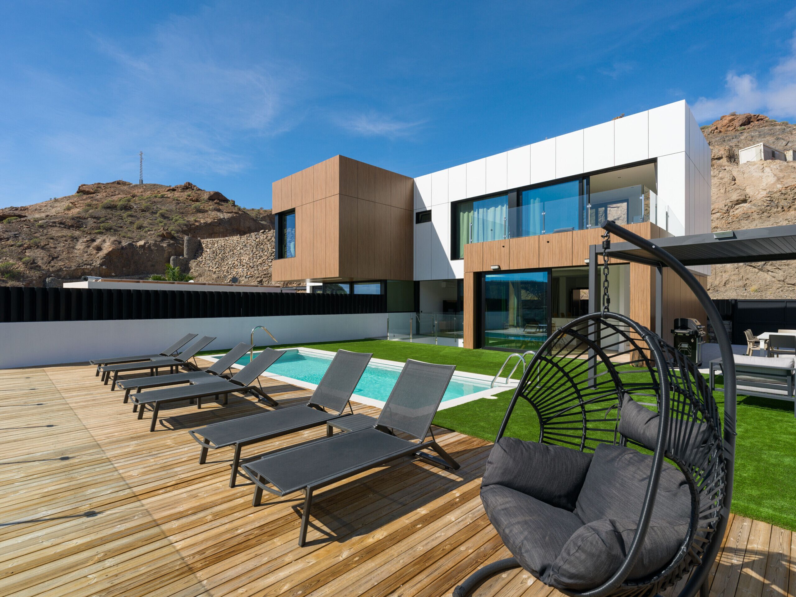 Un espace piscine extérieur moderne, doté de chaises longues et d’un fauteuil suspendu, entouré d’une terrasse en bois et d’un bâtiment aux grandes fenêtres.