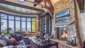 A spacious living room with a stone fireplace, large windows overlooking a body of water, and a cozy seating area. - Tewesi Manor | Historic Tahoe Lakefront w/ Private Pier (Carnelian Bay)