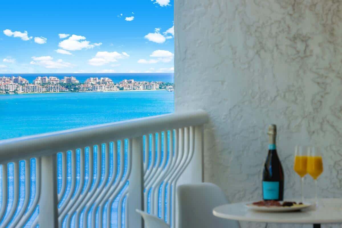 A balcony with a view of a coastal city, a table with a bottle and glasses, and a white chair.