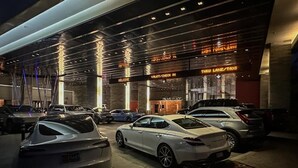 Ein modernes Gebäude mit mehreren Eingängen, auf denen "Welcome", "Valet/Check In" und "Thru Lane/Taxi" stehen. Mehrere Luxusautos sind vor dem Gebäude geparkt.