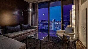 Ein modernes Hotelzimmer mit Sofa, Sessel und Glastisch. Ein großes Fenster bietet Blick auf die Stadt bei Nacht.