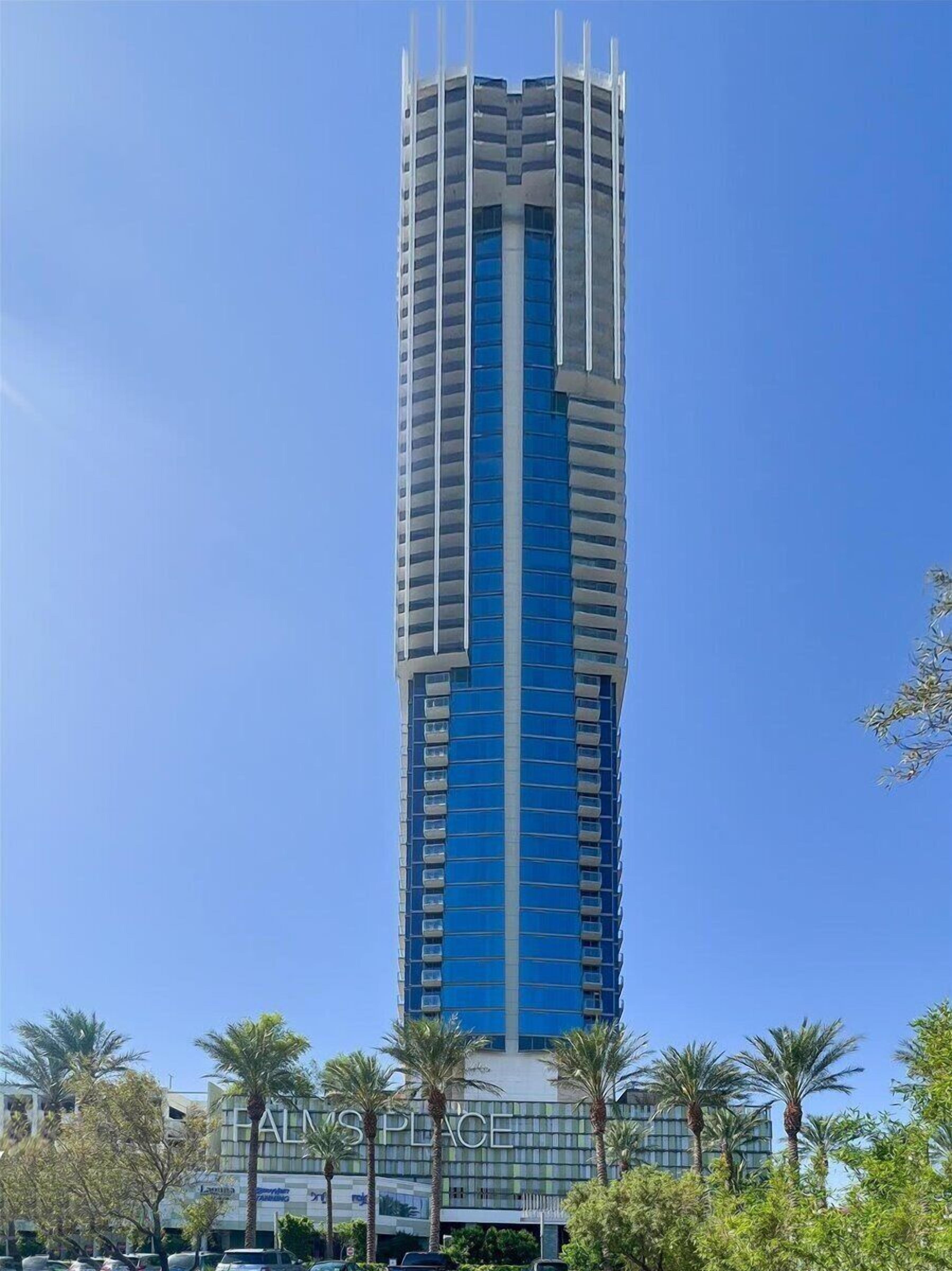 Een hoog, modern gebouw met een blauwe gevel en witte accenten, omringd door palmbomen.
