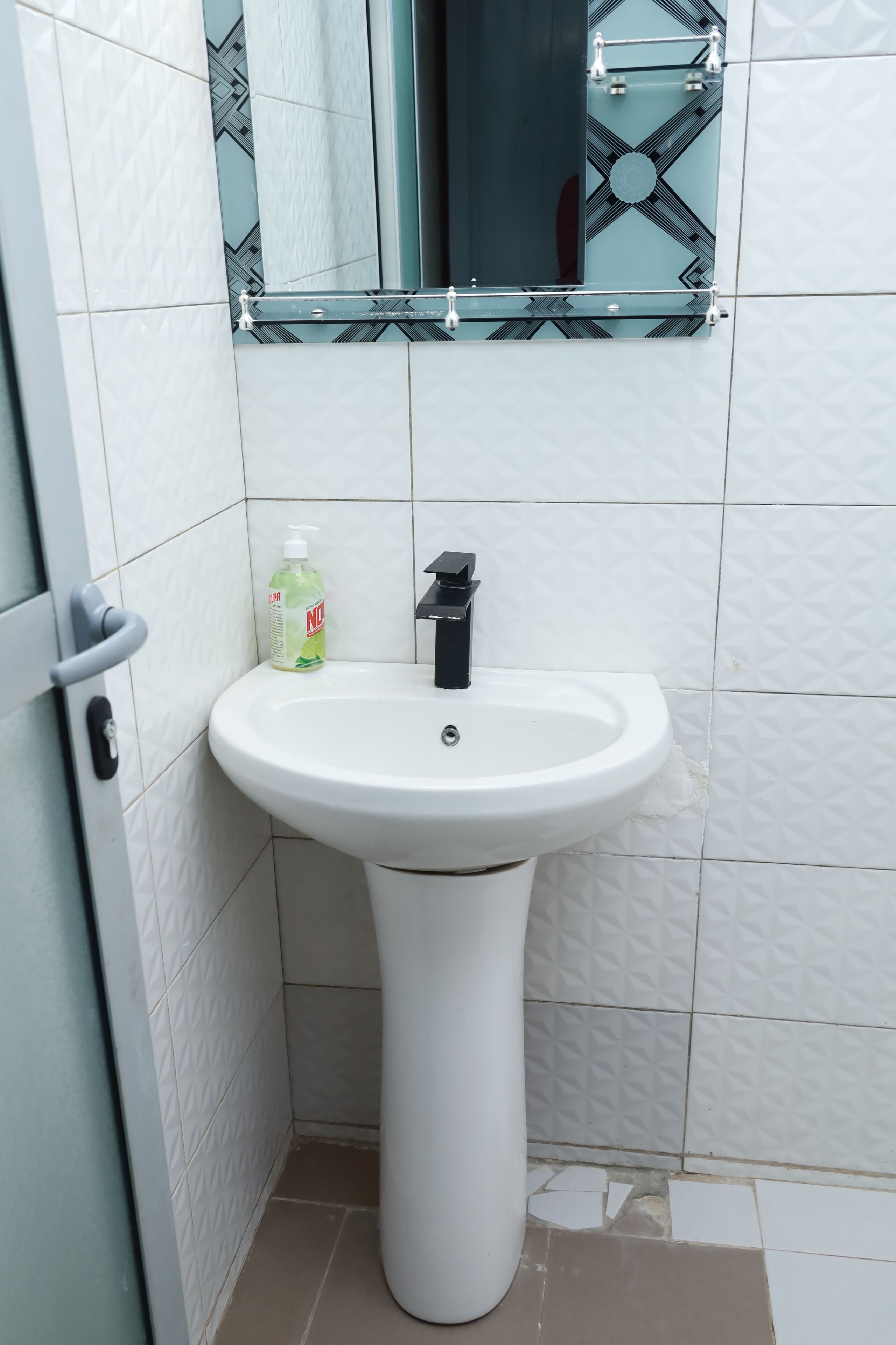 A white pedestal sink with a black faucet, a bottle of hand soap, and a mirror with a geometric pattern above it.