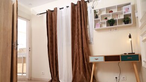 Una stanza con una porta in legno, una finestra vista, una scrivania con cassetti, uno scaffale con piante e una presa elettrica.