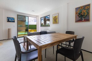 A dining area with a wooden table and black chairs, a painting of sunflowers on the wall, and a view of a balcony with a yellow table.